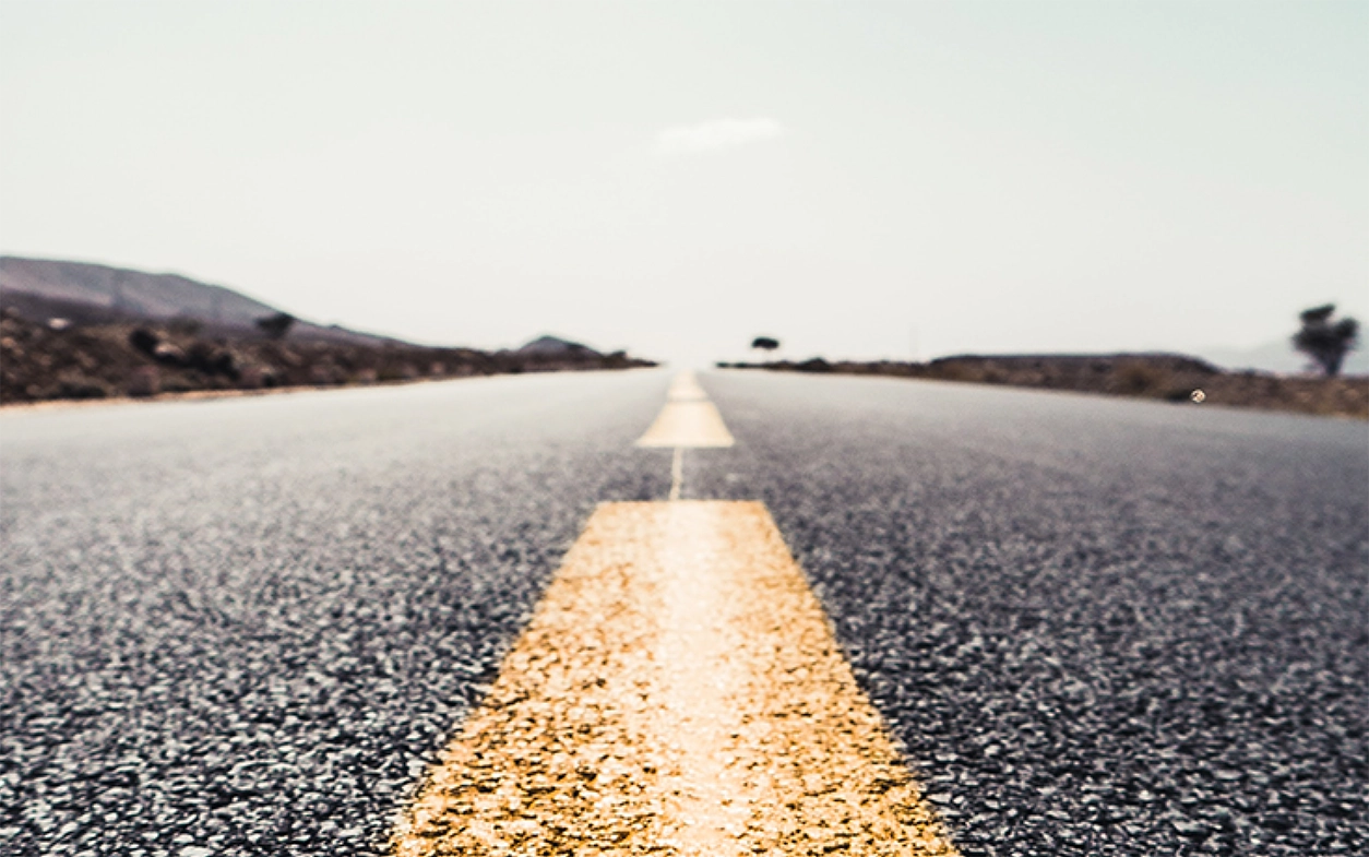 A close up shot of an asphalt road with a bright yellow painted, dashed divider line going straight down the middle toward the horizon. This represents that adaptive resilience has no finish line.