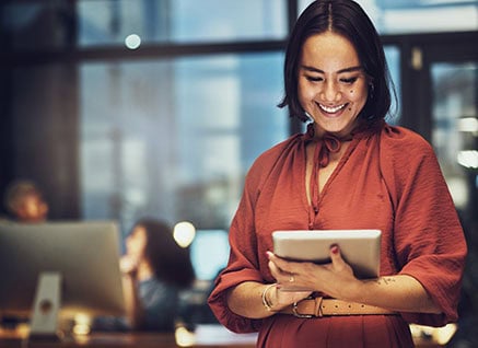 A young Asian woman in an office, wearing a terracotta-colored blouse, smiling down at a tablet. She represents what brand measurement with purpose looks like,